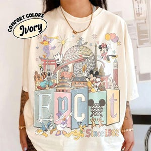 May include: An ivory t-shirt featuring a colorful graphic print of various cartoon characters, the Epcot geodesic sphere, and landmarks. The text reads Epcot and Since 1982. A Comfort Colors Ivory label is visible in the upper left corner of the image.