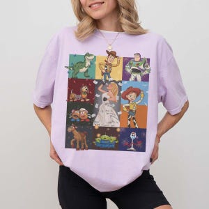 May include: A light purple short-sleeve t-shirt featuring a grid of nine colorful illustrations of various animated characters, including a green dinosaur, a cowboy, a space ranger, a dog, a singer, a cowgirl, potato figures, a horse, aliens, and a fork.