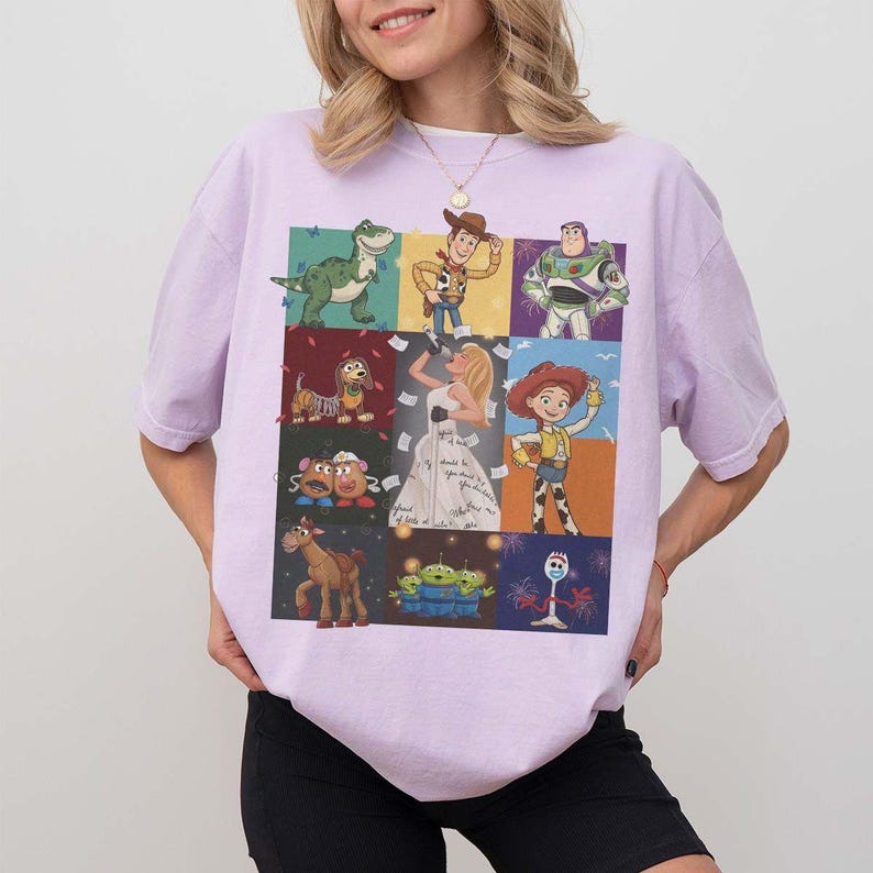 May include: A light purple short-sleeve t-shirt featuring a grid of nine colorful illustrations of various animated characters, including a green dinosaur, a cowboy, a space ranger, a dog, a singer, a cowgirl, potato figures, a horse, aliens, and a fork.