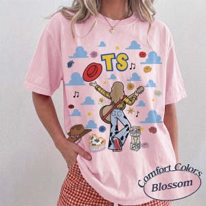 May include: A light pink short-sleeve t-shirt featuring a graphic print of a person playing a guitar, a red hat, blue clouds, flowers, and the text TS. The design includes a stack of blocks with the letters T and S and a cow print pattern.