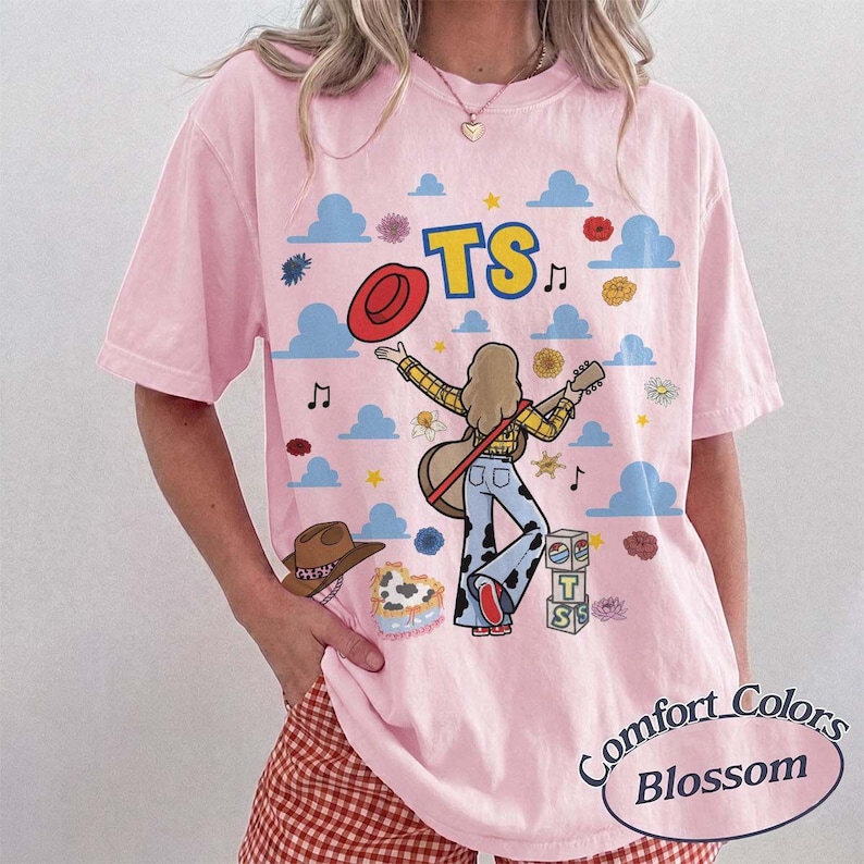 May include: A light pink short-sleeve t-shirt featuring a graphic print of a person playing a guitar, a red hat, blue clouds, flowers, and the text TS. The design includes a stack of blocks with the letters T and S and a cow print pattern.