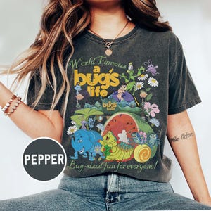 May include: Dark gray t-shirt featuring a colorful graphic from "A Bug's Life." The design includes the text "World Famous a bug's life" and "Bug-sized fun for everyone!" with cartoon bugs, flowers, and a watermelon.