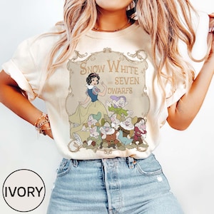 May include: Ivory-colored t-shirt featuring a graphic of Snow White and the Seven Dwarfs. The design includes an illustration of Snow White in a yellow dress and the seven dwarfs. The text "Snow White and Seven Dwarfs" is displayed above the characters.