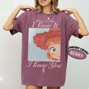 May include: A berry-colored short-sleeve t-shirt featuring a graphic of a red-haired character wearing a red hat. The text reads EST 2026 I Knew It I Knew You with a small musical note symbol. A circular label on the right side reads COMFORT COLORS BERRY.