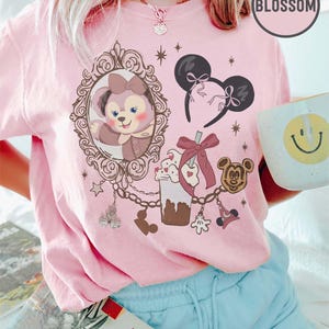 Könnte beinhalten: Hellrosa T-Shirt mit Disney-Motiven, darunter eine Cartoonfigur in einem Rahmen, Mickey Mouse Ohren und ein Milchshake. Das Wort "Blossom" ist auf dem T-Shirt aufgedruckt.