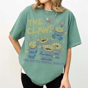May include: Green t-shirt featuring the phrase "THE CLAW" in yellow-green, with cartoon alien characters in blue suits. The text "THE MYSTIC PORTAL AWAITS" is below the aliens. The shirt has a relaxed fit.