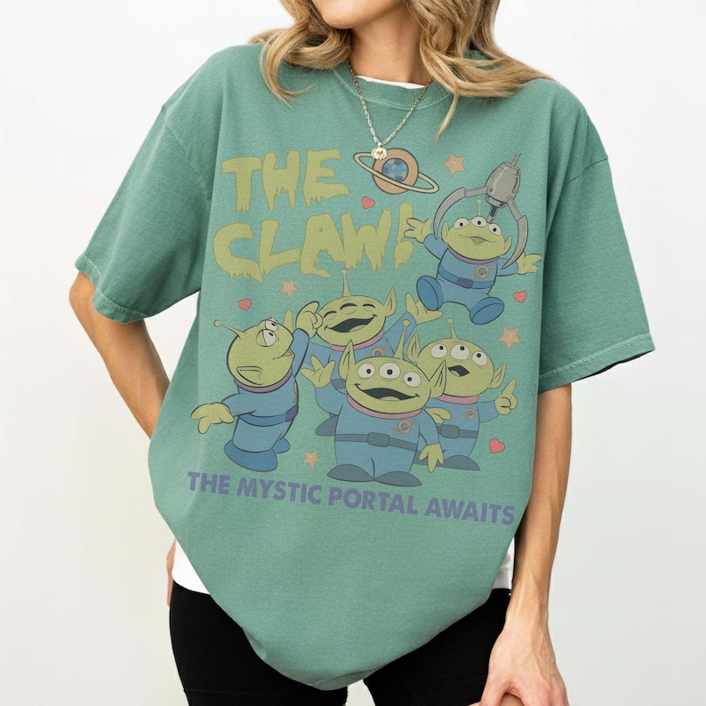 May include: Green t-shirt featuring the phrase "THE CLAW" in yellow-green, with cartoon alien characters in blue suits. The text "THE MYSTIC PORTAL AWAITS" is below the aliens. The shirt has a relaxed fit.