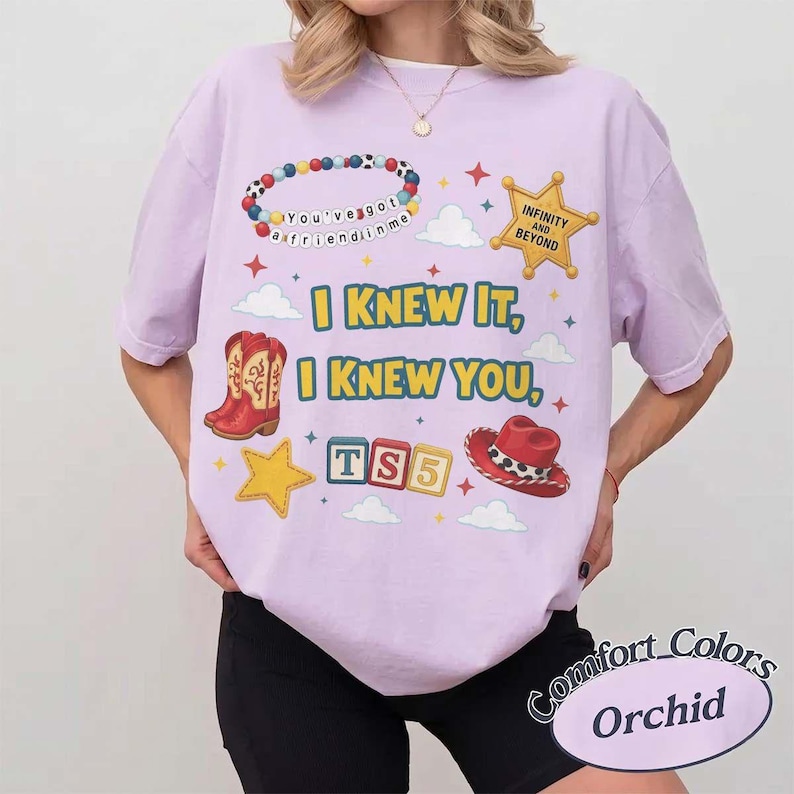 May include: A light purple t-shirt featuring a graphic print with a beaded bracelet that reads You've got a friend in me, a yellow sheriff star with Infinity and Beyond, red cowboy boots, a red hat, and blocks spelling TS5 with the text I KNEW IT, I KNEW YOU.