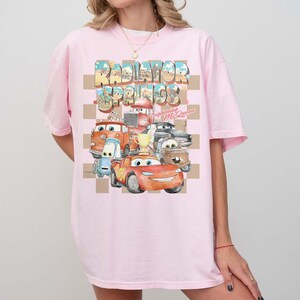 Peut inclure: T-shirt rose clair avec un motif color&eacute; de personnages du film "Cars". Le design comprend le texte "Radiator Springs" et "Lightning McQueen" dans un style vintage, sur un fond &agrave; carreaux.