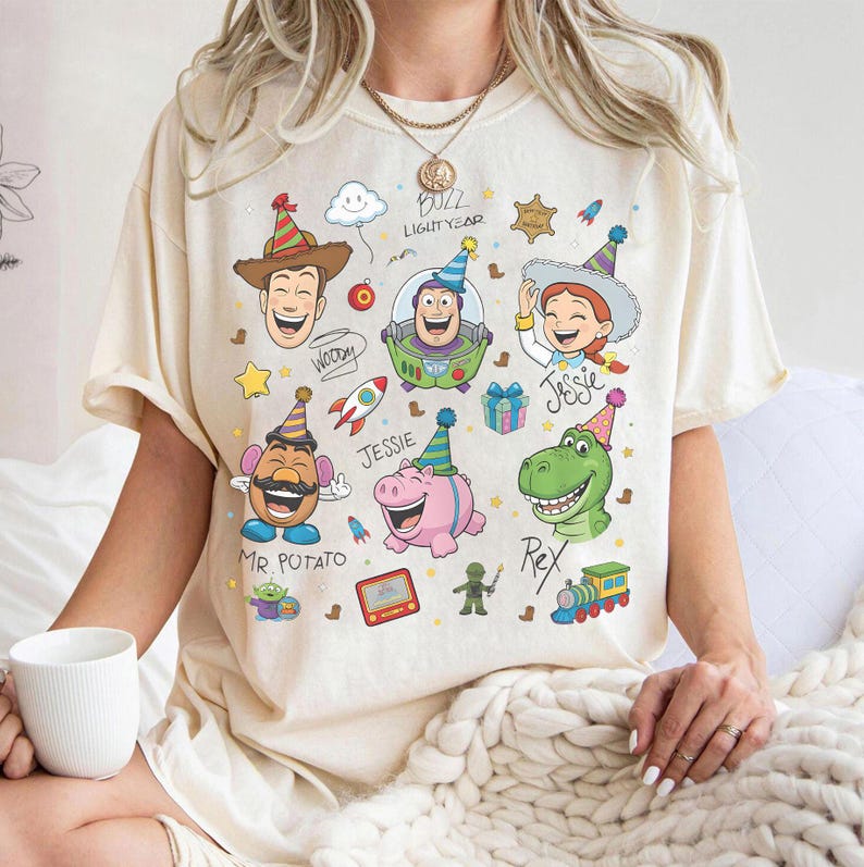 May include: Cream-colored t-shirt featuring colorful cartoon illustrations of characters from the Toy Story movie. The design includes Woody, Buzz Lightyear, Jessie, Mr. Potato Head, Rex, and Hamm, with the text "Buzz Lightyear".