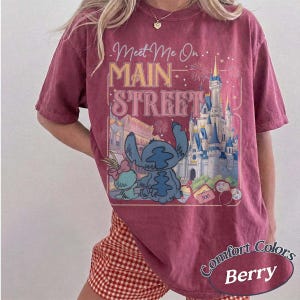 May include: Berry-colored t-shirt with a graphic featuring Stitch and Angel in front of a castle, with the text "Meet Me On Main Street." The shirt has a vintage, washed look, and the words "Comfort Colors" and "Berry" are also visible.