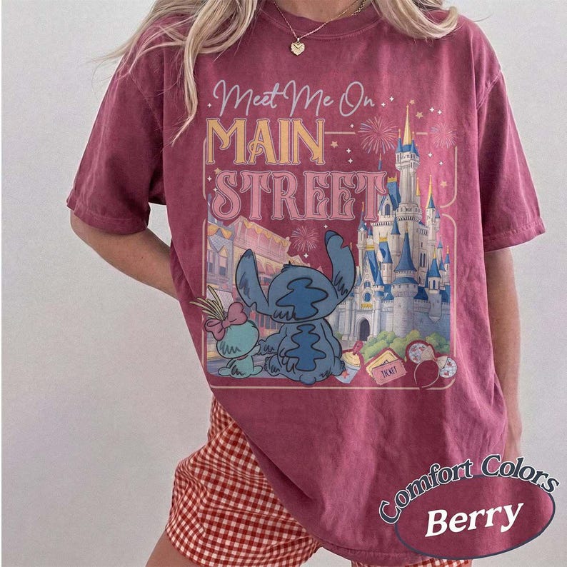 May include: Berry-colored t-shirt with a graphic featuring Stitch and Angel in front of a castle, with the text "Meet Me On Main Street." The shirt has a vintage, washed look, and the words "Comfort Colors" and "Berry" are also visible.