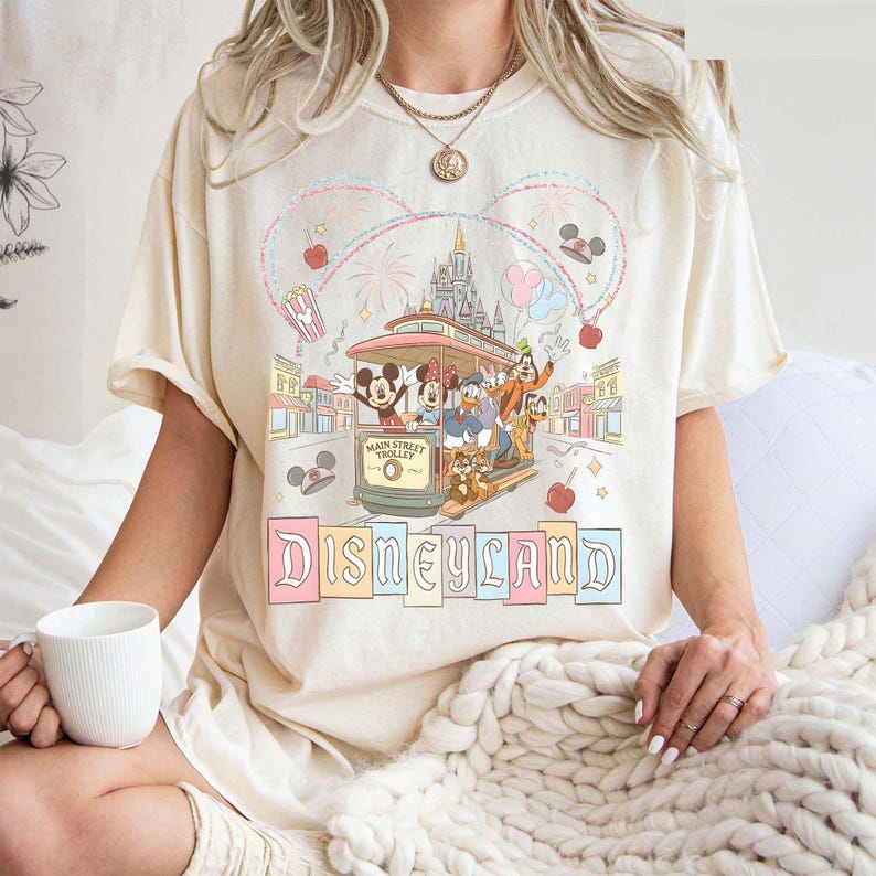 May include: Cream-colored t-shirt featuring a Disneyland design. The graphic shows Mickey Mouse, Minnie Mouse, Donald Duck, and Goofy riding a Main Street Trolley. The word "Disneyland" is written in pastel colors below the trolley.
