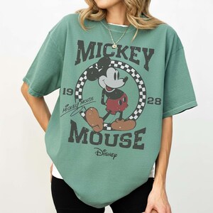 May include: Green t-shirt featuring Mickey Mouse. The graphic includes Mickey Mouse in a circle with a checkered pattern, the words "Mickey Mouse", "1928", and "Disney". The shirt has short sleeves and a crew neck.