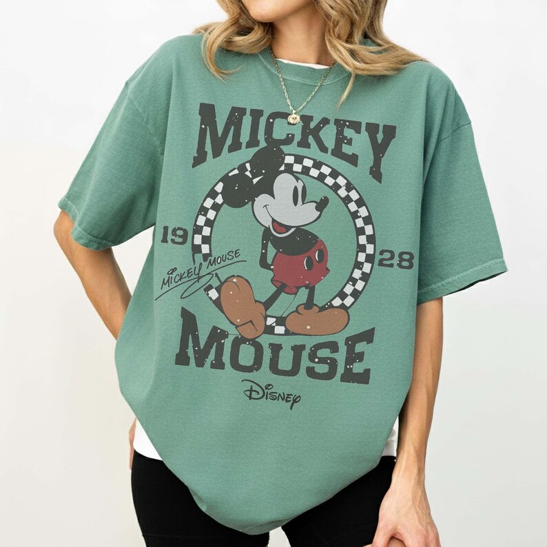May include: Green t-shirt featuring Mickey Mouse. The graphic includes Mickey Mouse in a circle with a checkered pattern, the words "Mickey Mouse", "1928", and "Disney". The shirt has short sleeves and a crew neck.