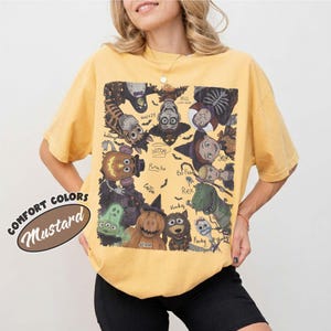 May include: A mustard yellow short-sleeve t-shirt featuring a large rectangular graphic print of various animated characters in Halloween costumes. The design includes text labels for characters like Woody, Buzz, Rex, and Forky, arranged in a circular pattern.