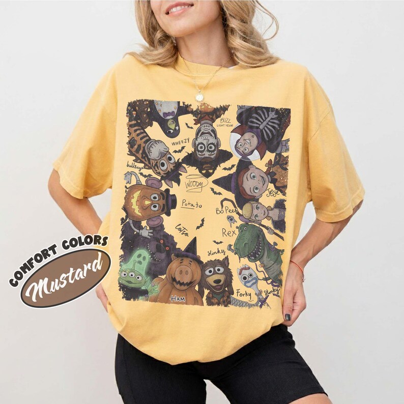 May include: A mustard yellow short-sleeve t-shirt featuring a large rectangular graphic print of various animated characters in Halloween costumes. The design includes text labels for characters like Woody, Buzz, Rex, and Forky, arranged in a circular pattern.