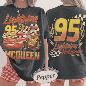 Może przedstawiać: Ciemnoszara koszulka z Lightning McQueen z filmu Auta. Prz&oacute;d przedstawia McQueena z numerem 95, flagą w szachownicę i napisem "Lightning McQueen". Tył ma duży numer 95 z płomieniami i "Lightning McQueen".