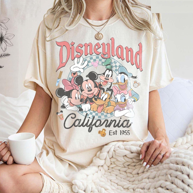 May include: Cream-colored t-shirt featuring a Disneyland California graphic with Mickey Mouse, Minnie Mouse, Donald Duck, Goofy, and Pluto. The graphic is in a circle with a pink and blue checkered background. The text "Est 1955" is below the word "California".