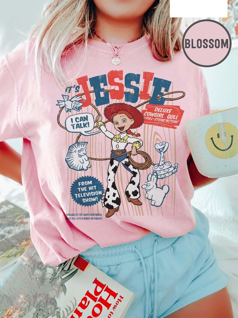 May include: Pale pink t-shirt with a graphic of Jessie from "Toy Story." The design includes the text "It's Jessie," "Deluxe Cowgirl Doll," and cartoon characters. The shirt also has the text "I can talk!" and "From the hit television show!"