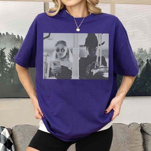 May include: A purple t-shirt featuring a black and white image of two people. One person is wearing sunglasses and holding a phone, and the other is wearing a hat. The shirt is a solid color and has short sleeves.