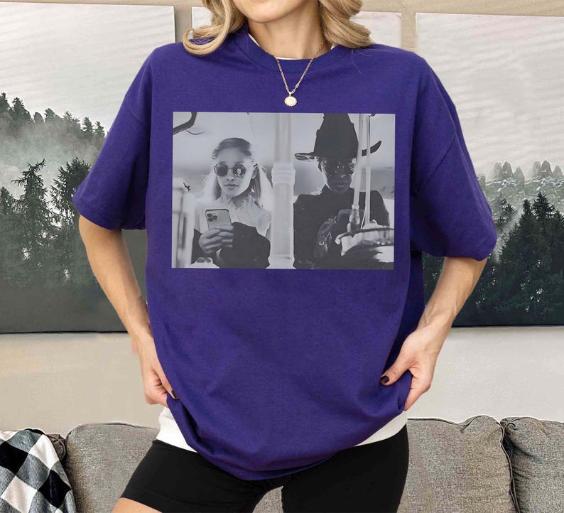 May include: A purple t-shirt featuring a black and white image of two people. One person is wearing sunglasses and holding a phone, and the other is wearing a hat. The shirt is a solid color and has short sleeves.
