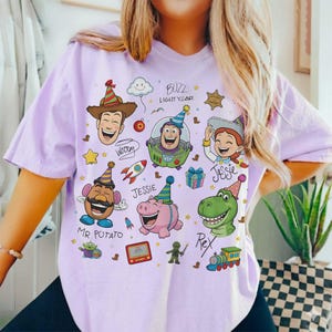 May include: A light purple t-shirt featuring cartoon characters from the movie Toy Story. The design includes Woody, Buzz Lightyear, Jessie, Mr. Potato Head, Rex, and Hamm, with party hats and other festive elements.