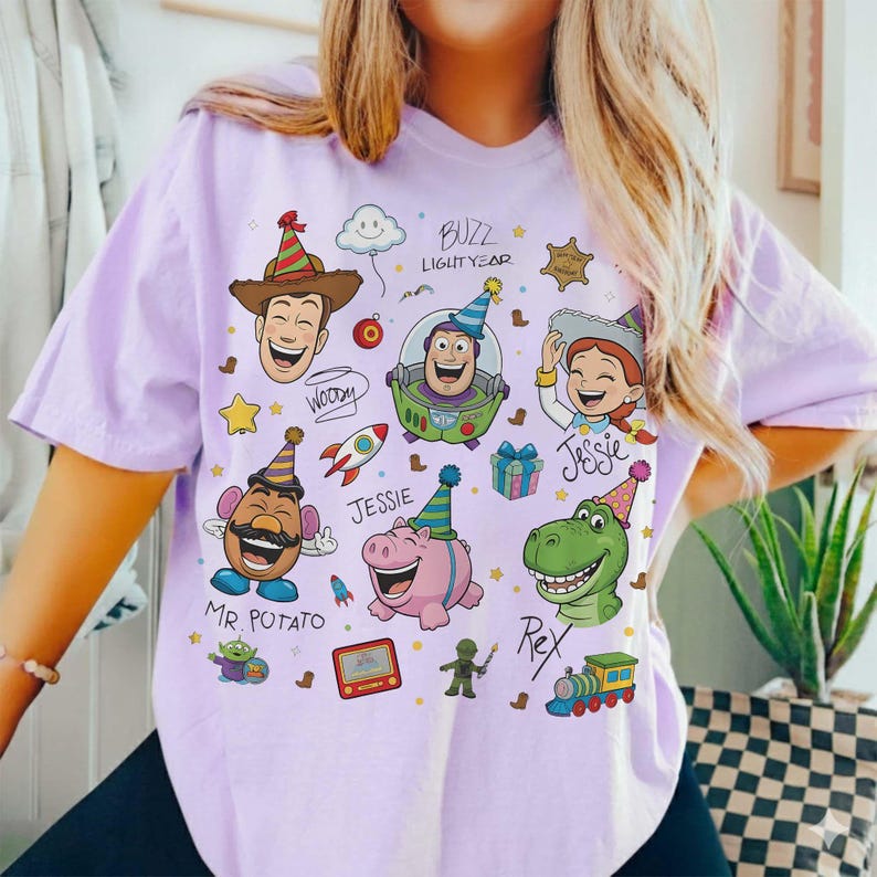 May include: A light purple t-shirt featuring cartoon characters from the movie Toy Story. The design includes Woody, Buzz Lightyear, Jessie, Mr. Potato Head, Rex, and Hamm, with party hats and other festive elements.