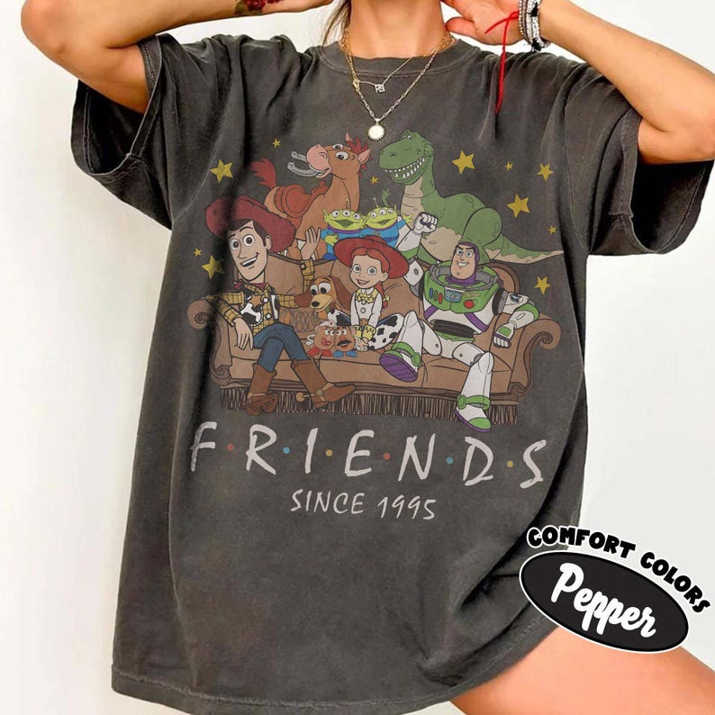 May include: A charcoal gray short-sleeve t-shirt featuring a graphic print of Toy Story characters sitting on a couch. The text reads "FRIENDS SINCE 1995" below the illustration. A small black oval label in the corner says "COMFORT COLORS Pepper".