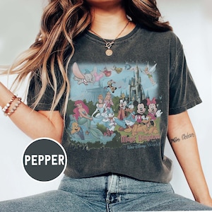 K&ouml;nnte beinhalten: Dunkelgraues T-Shirt mit einer farbenfrohen Grafik von Disney-Figuren, darunter Micky Maus, Arielle und Cinderella, vor einem Schloss. Der Text "Magic Kingdom" und "Walt Disney World" ist darunter gedruckt.