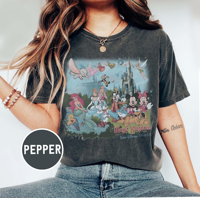 K&ouml;nnte beinhalten: Dunkelgraues T-Shirt mit einer farbenfrohen Grafik von Disney-Figuren, darunter Micky Maus, Arielle und Cinderella, vor einem Schloss. Der Text "Magic Kingdom" und "Walt Disney World" ist darunter gedruckt.