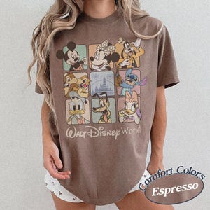 May include: Espresso-colored t-shirt featuring a grid of Disney characters, including Mickey Mouse, Minnie Mouse, and Goofy. The shirt has the words "Walt Disney World" printed below the grid. The shirt is a Comfort Colors brand.