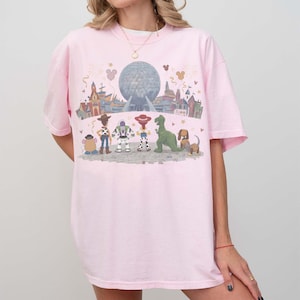 Puede incluir: Camiseta rosa claro con un gr&aacute;fico colorido de personajes de Disney y una escena de parque. El dise&ntilde;o incluye a Woody, Buzz Lightyear, Rex y otros personajes. La camiseta tiene un ajuste c&oacute;modo y holgado.