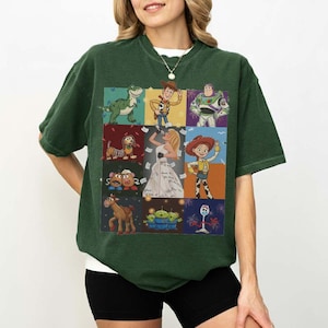 May include: A dark green short-sleeve t-shirt featuring a grid of nine colorful illustrations of various animated characters, including a green dinosaur, a cowboy, a space ranger, a dog, a blonde singer, a cowgirl, potato figures, a horse, and aliens.