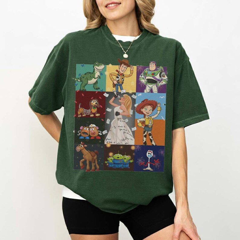 May include: A dark green short-sleeve t-shirt featuring a grid of nine colorful illustrations of various animated characters, including a green dinosaur, a cowboy, a space ranger, a dog, a blonde singer, a cowgirl, potato figures, a horse, and aliens.