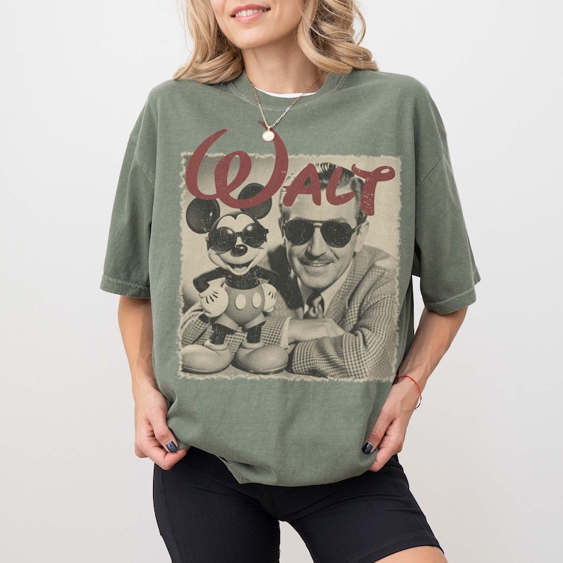 Puede incluir: Camiseta verde oliva con un gr&aacute;fico vintage de Mickey Mouse y Walt Disney. El gr&aacute;fico incluye la palabra "WALT" en una fuente roja estilizada. Mickey Mouse lleva gafas de sol. La camiseta tiene un corte relajado.