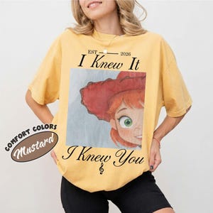 May include: A mustard yellow short-sleeve t-shirt featuring a graphic print of a red-haired character wearing a red hat. The text reads "EST 2026 I Knew It" above the graphic and "I Knew You" below it. A separate circular graphic reads "COMFORT COLORS MUSTARD".