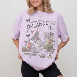 K&ouml;nnte beinhalten: Ein helllila T-Shirt mit einer Grafik von Orlando, Florida. Das Design enth&auml;lt den Text "Greetings from Orlando, FL" und Illustrationen eines Schlosses, Feuerwerks und anderer Attraktionen. Das Shirt ist ein bequemer, l&auml;ssiger Stil.