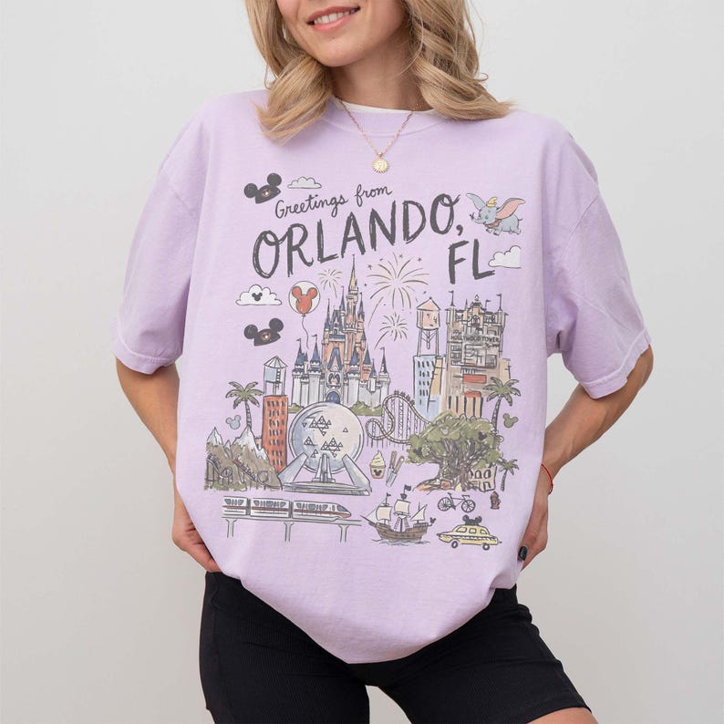 K&ouml;nnte beinhalten: Ein helllila T-Shirt mit einer Grafik von Orlando, Florida. Das Design enth&auml;lt den Text "Greetings from Orlando, FL" und Illustrationen eines Schlosses, Feuerwerks und anderer Attraktionen. Das Shirt ist ein bequemer, l&auml;ssiger Stil.