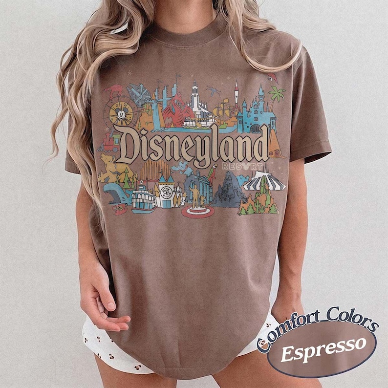May include: Brown t-shirt with a colorful Disneyland Resort graphic. The design features various park attractions and the Disneyland Resort logo. The shirt is labeled "Comfort Colors Espresso".