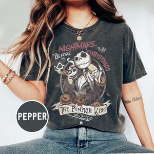 May include: Dark gray t-shirt featuring a graphic from "The Nightmare Before Christmas." The design includes Jack Skellington, pumpkins, and the text "The Pumpkin King." The shirt has a vintage, worn look.