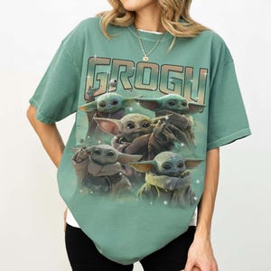 Puede incluir: Una camiseta verde con la palabra "GROGU" en la parte superior en un estilo retro. Debajo del texto hay un gr&aacute;fico de Grogu, tambi&eacute;n conocido como Baby Yoda, de The Mandalorian. El gr&aacute;fico muestra m&uacute;ltiples im&aacute;genes de Grogu. La camiseta tiene mangas cortas.