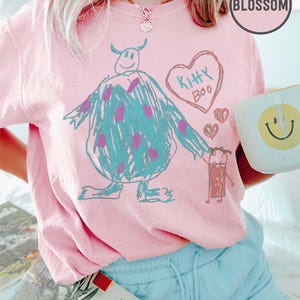 May include: A light pink t-shirt featuring a hand-drawn cartoon of a blue monster with purple spots, a heart with the words "Kitty Boo," and a small figure. The shirt is paired with light blue shorts and a mug with a yellow smiley face.