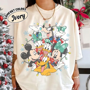 May include: Ivory Comfort Colors t-shirt with a Christmas design. It features Disney characters Mickey, Minnie, Donald, Daisy, Goofy, and Pluto, surrounded by fairy lights and snowflakes.