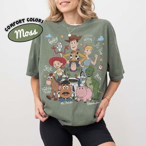 May include: A moss green short-sleeve t-shirt featuring a graphic print of Toy Story characters including Woody, Buzz Lightyear, Jessie, Bullseye, Mr. Potato Head, Hamm, Forky, and Bo Peep, with their names written in a white script font.