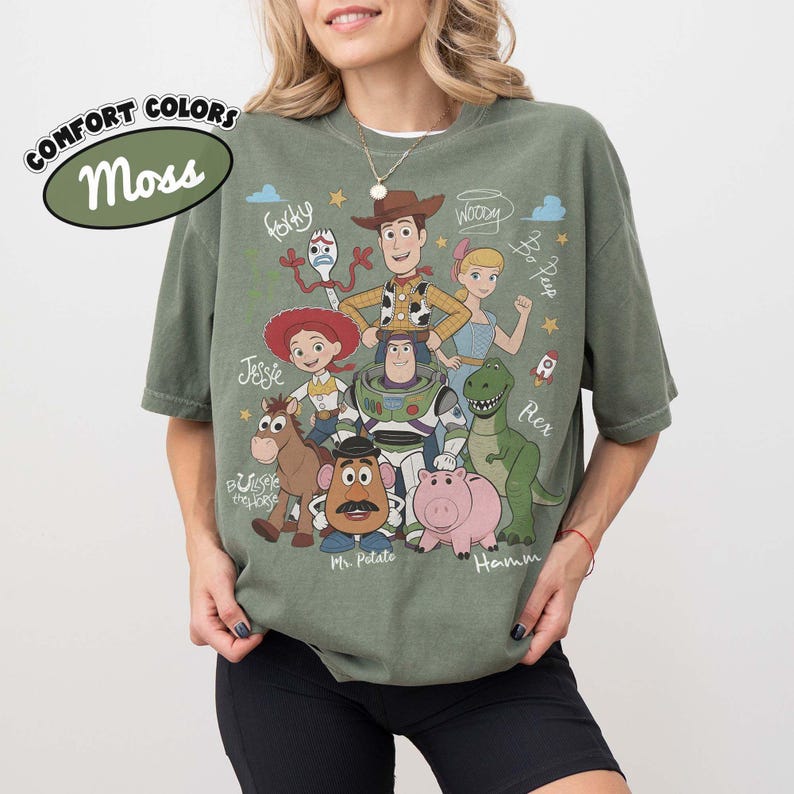 May include: A moss green short-sleeve t-shirt featuring a graphic print of Toy Story characters including Woody, Buzz Lightyear, Jessie, Bullseye, Mr. Potato Head, Hamm, Forky, and Bo Peep, with their names written in a white script font.