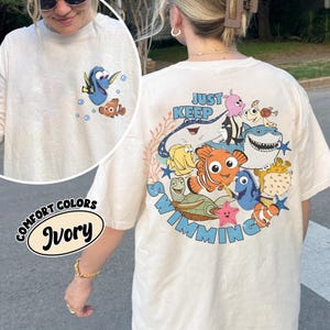 May include: Ivory-colored long-sleeve t-shirt featuring a cartoon image of Dory and Nemo on the front. The back of the shirt displays a larger graphic with the phrase "Just Keep Swimming" and various sea creatures. The shirt is part of the Comfort Colors collection.