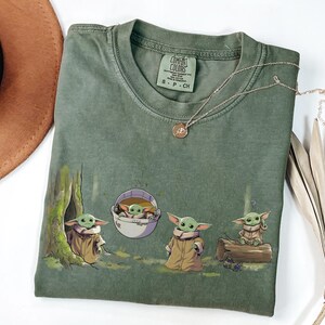 May include: An olive green short-sleeve t-shirt featuring a graphic print of four small green creatures with large ears and tan robes. One creature sits in a tree hollow, one in a floating pod, one stands, and one sits on a log. The shirt has a crew neck.