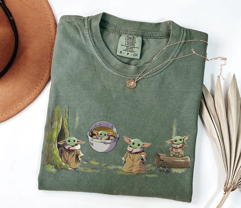 May include: An olive green short-sleeve t-shirt featuring a graphic print of four small green creatures with large ears and tan robes. One creature sits in a tree hollow, one in a floating pod, one stands, and one sits on a log. The shirt has a crew neck.