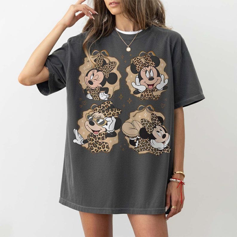 May include: Dark gray t-shirt featuring four images of Minnie Mouse in leopard print outfits. Each image is set within a tan oval with gold star accents. Minnie is wearing a bow and various poses. The shirt is a casual, oversized style.
